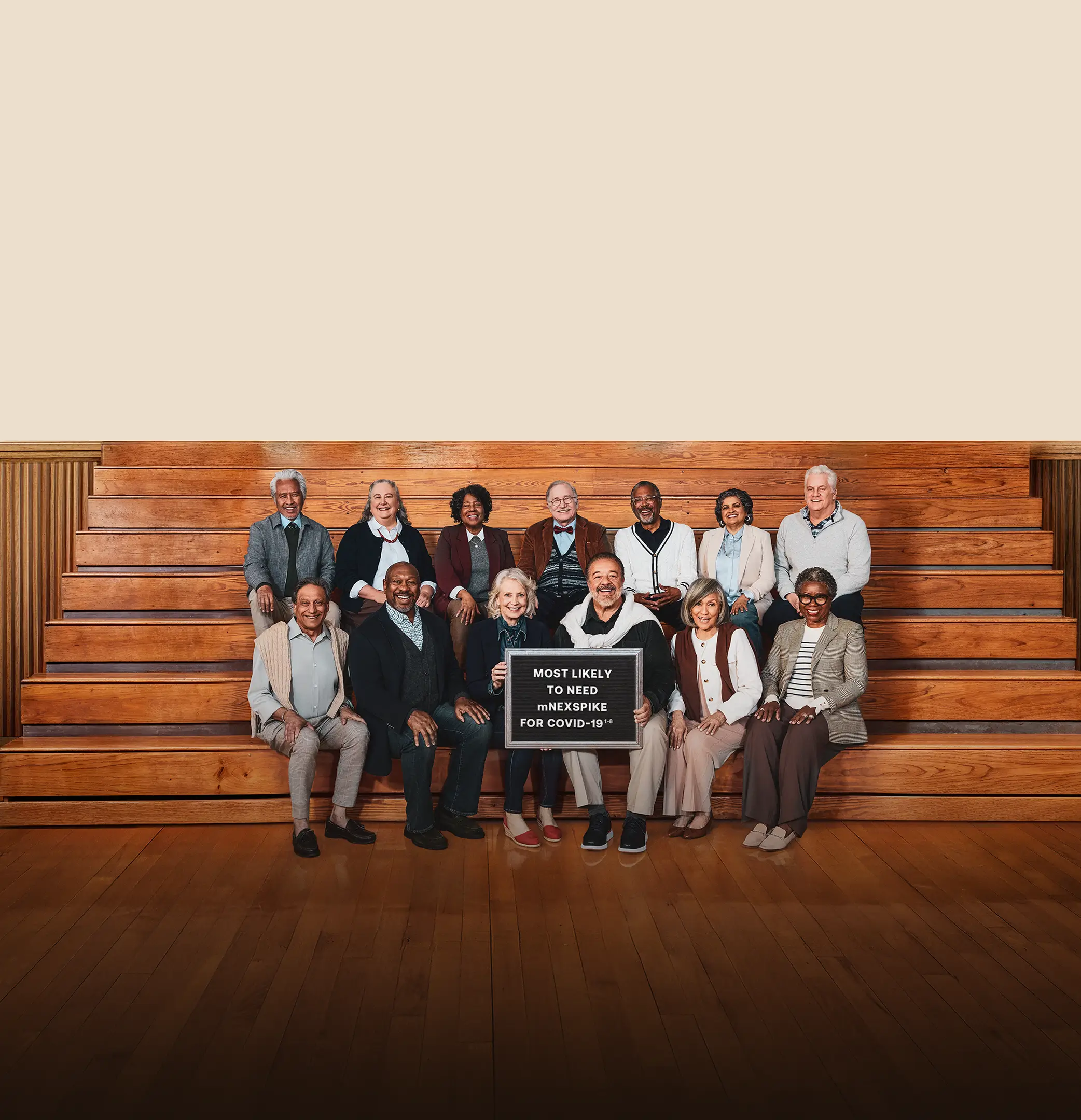 A group of older adults smiling and posing for a photo on gym bleachers. A green banner reads “Here for the Senior Class.” Text beside the banner promotes the mNEXSPIKE® COVID-19 vaccine for people aged 65 and older. One person in the front row holds a sign that says, “Most likely to need mNEXSPIKE for COVID-19."
