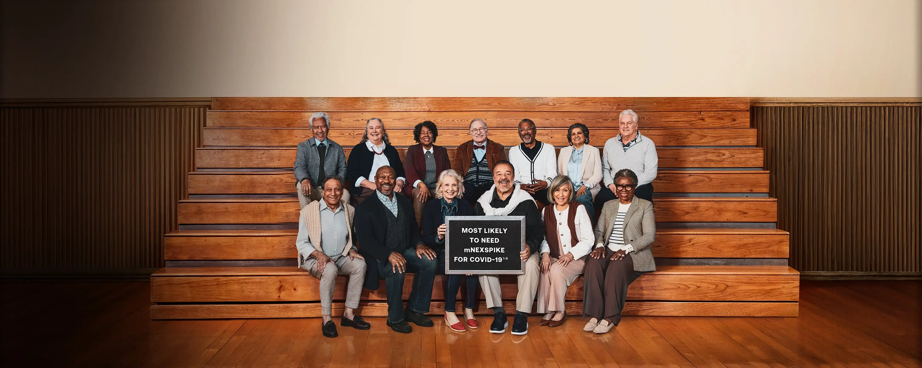 A group of older adults smiling and posing for a photo on gym bleachers. A green banner reads “Here for the Senior Class.” Text beside the banner promotes the mNEXSPIKE® COVID-19 vaccine for people aged 65 and older. One person in the front row holds a sign that says, “Most likely to need mNEXSPIKE for COVID-19."